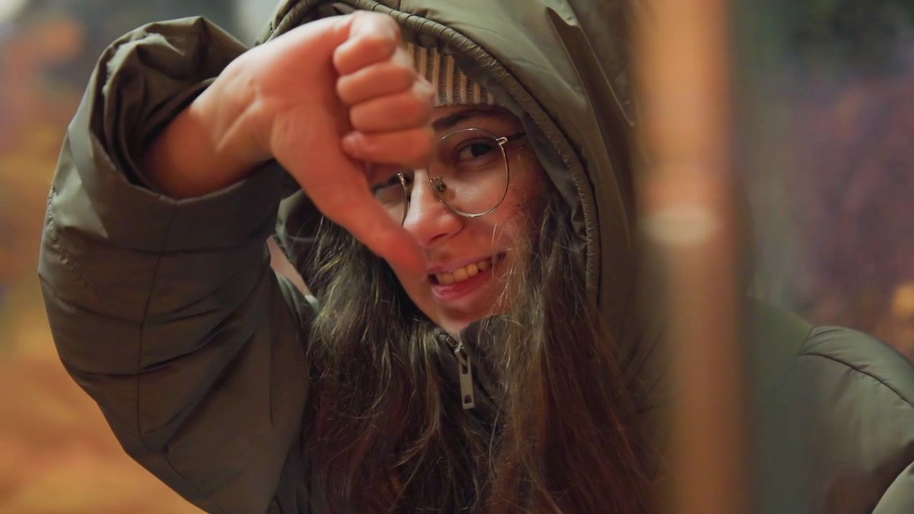 Jonge vrouw met capuchon steekt duim omlaag in een nachtelijke bushalte, draagt een muts en bril, afwijzend gebaar. Neon bokeh lichten, subtiele frons, korrelige filmtextuur, koude jas, openhartige kritische sfeer.