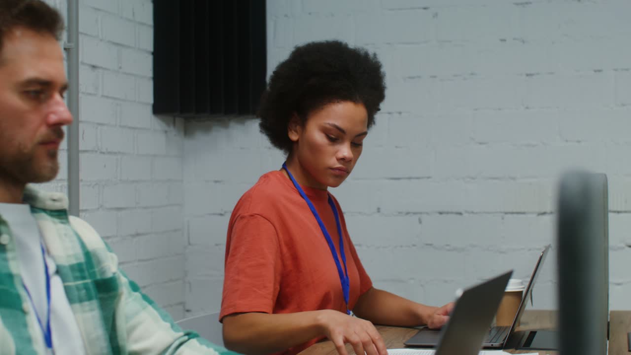 A man and woman work at laptops, sitting at one work desk in a office