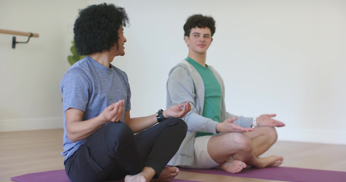 African American hooded men meditating on purple mats in room, left man turning gaze, smiling