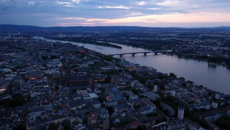 Alta-Toma-Aérea-De-Drones-De-Maguncia-En-La-Hora-Mágica-De-La-Noche-Dando-Vueltas-Alrededor-Del-Centro-De-La-Ciudad-Con-La-Catedral-Y-El-Agua-Oscura-Del-Río-Rin-En-El-Fondo-Mostrando-Un-Cielo-Colorido