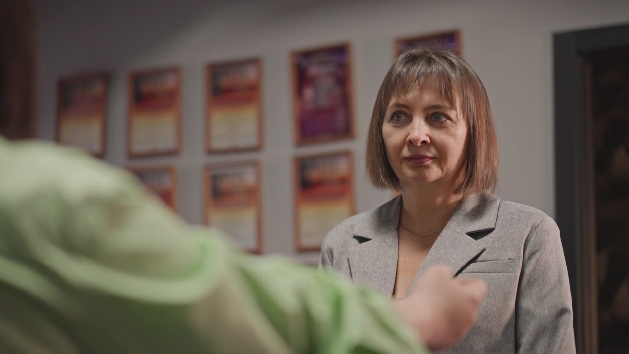 Direttore scolastico che consegna la lettera di accettazione a un candidato in un corridoio con poster. Una donna in blazer grigio sorride, porgendo documenti ufficiali, in un'atmosfera luminosa e incoraggiante, con segnaletica e display
