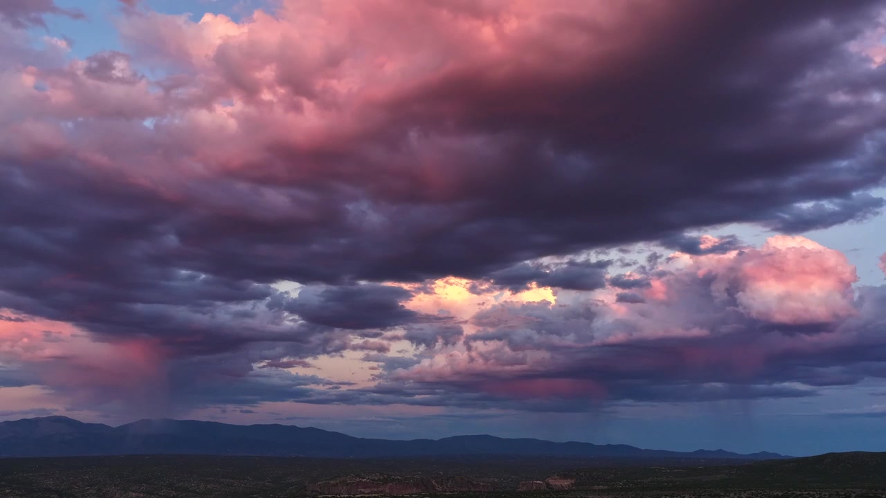 Hiperlapso aéreo 4K de nubes de lluvia tormentosas moviéndose sobre un cañón escarpado y un paisaje montañoso. El clima dramático cambia los colores del cielo de un vibrante púrpura y rosa a un gris oscuro y pesado.