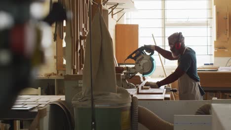 african american male carpenter cutting wooden plank with electric chop saw in a carpentry shop