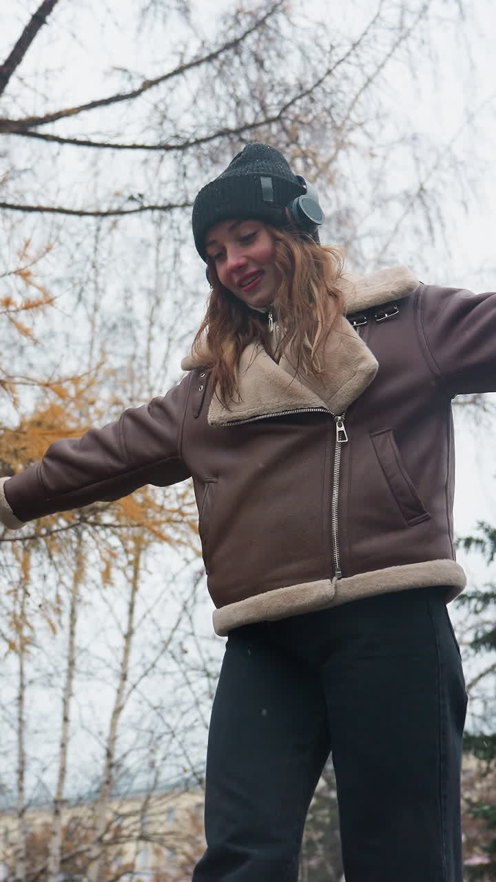 Happy girl wearing black knit cap brown shearling jacket black trousers balancing gracefully with arms outstretched while walking on wide stone surface in peaceful autumn park surrounded by c fallen leaves