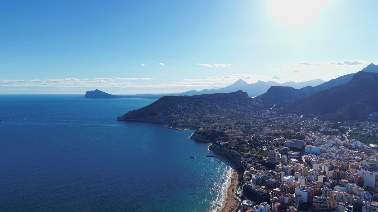 Costa di Calpe in Spagna che mostra acque turchesi del Mediterraneo, spiaggia sabbiosa, edifici della città costiera, promontorio roccioso e catene montuose sotto un cielo soleggiato e luminoso.