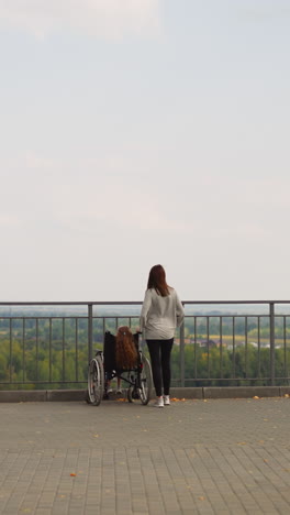 redhead woman and little girl in wheelchair enjoy scenic nature. mother and daughter with muscular weakness look at city park near river backside view