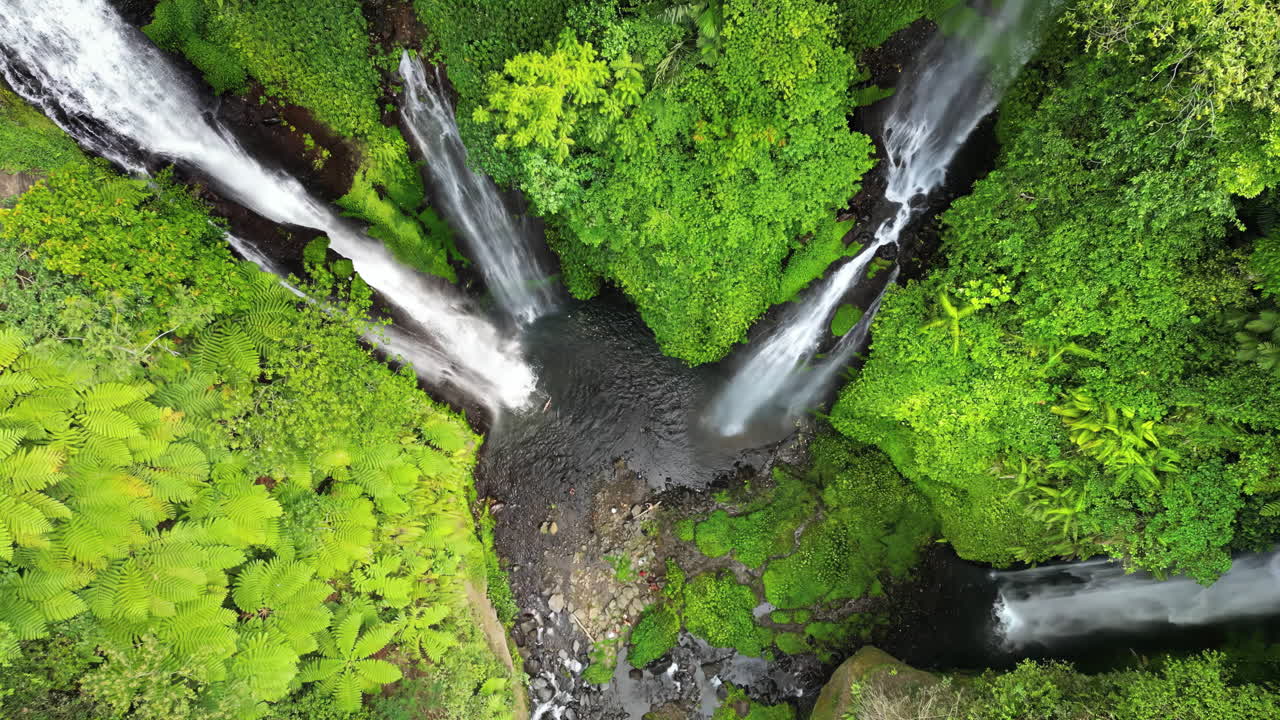 Pemandangan drone beberapa tingkatan Air Terjun Sekumpul di Bali Utara, Indonesia. Aliran air putih tinggi mengalir menuruni tebing hutan hujan curam yang tertutup vegetasi hijau subur.