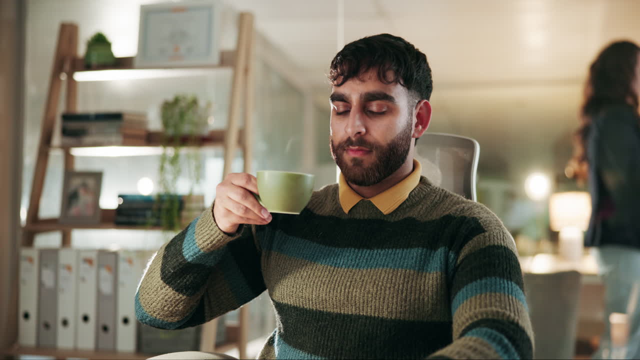 un hombre bebe café en su escritorio en una oficina mientras una mujer está trabajando en el fondo.