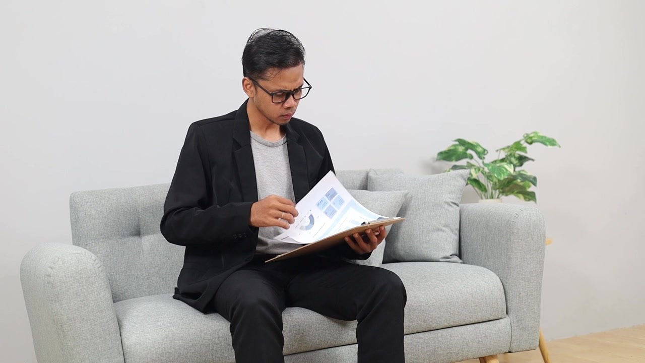 Serious Asian man freelancer work with documents on home couch reading information business papers prepare financial report