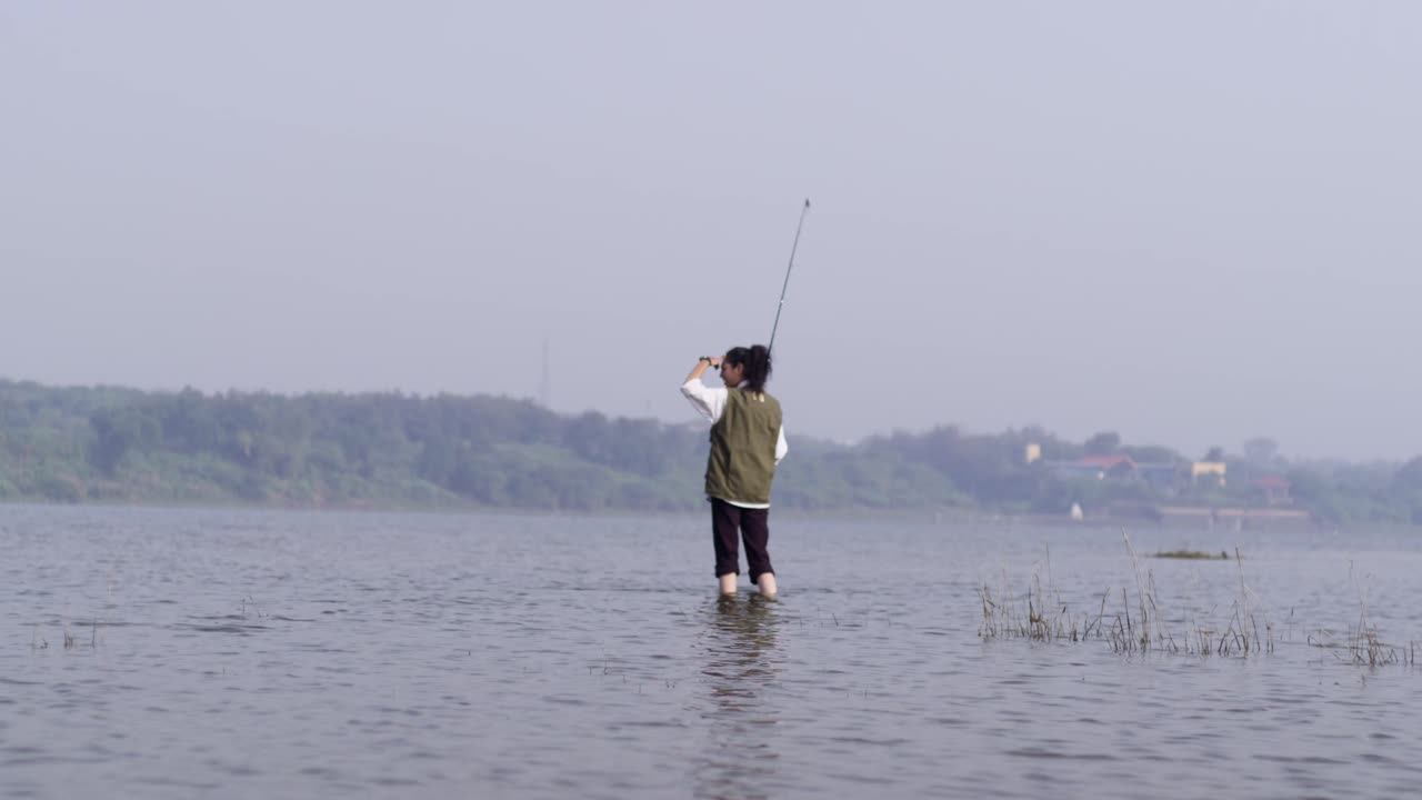 Seorang gadis Asia Selatan mengenakan rompi memancing sedang memancing di lanskap danau yang luas dikelilingi oleh tanaman hijau