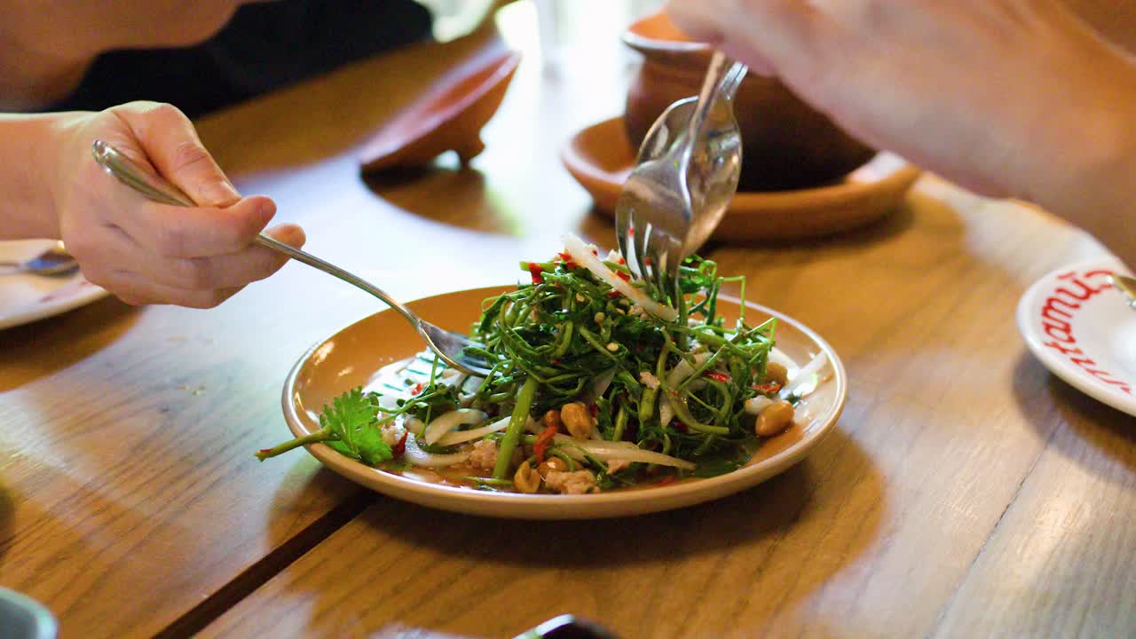Hands serve traditional spicy water mimosa salad at a well-lit Bangkok restaurant dining table