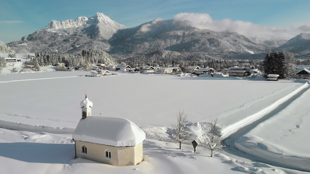 Aerial Drone shot showing a snowy landscape in in the sunshine of the Tyrol, Austria and a snow covered tiny chapel in Lechaschau