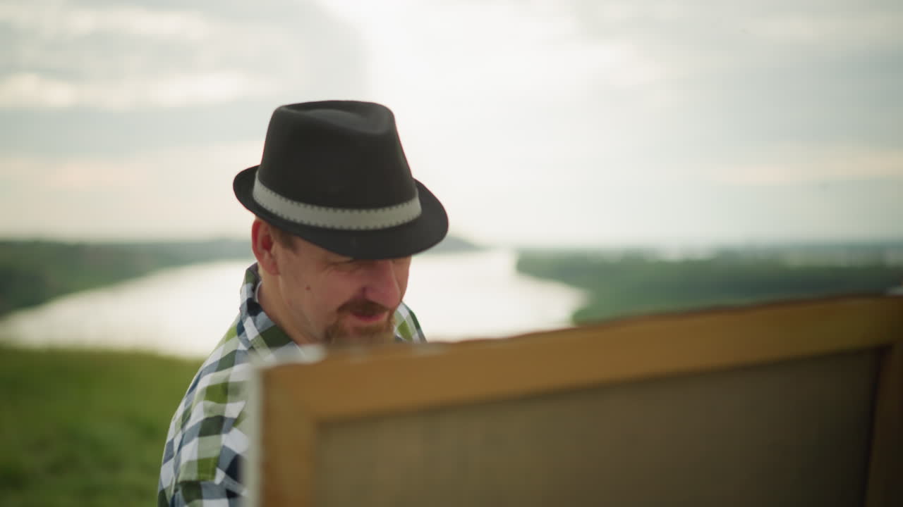 Close-up of a painter wearing a black hat, checkered shirt, and jeans, deeply focused on painting a large canvas set up in a tranquil grass field by a serene lake