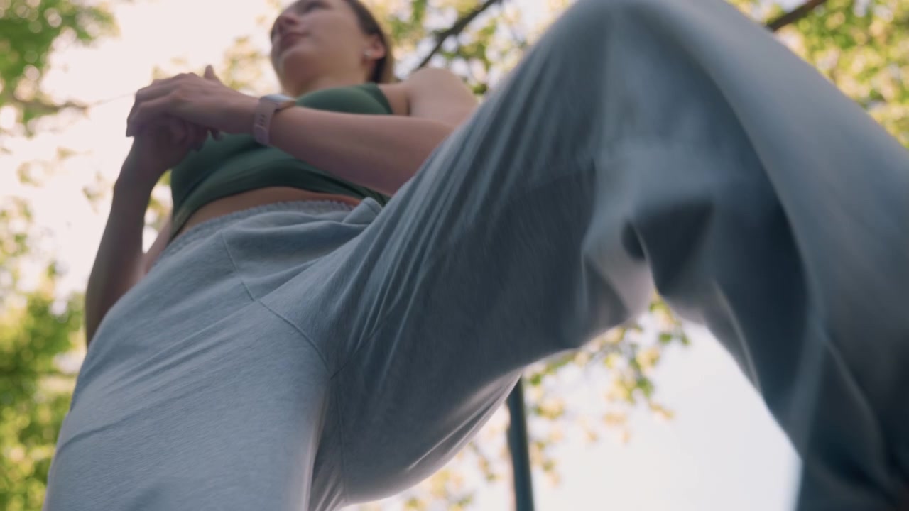 Close-up de jovem mulher realizando alongamentos de pernas ao ar livre à luz do dia, vestindo roupa esportiva casual e smartwatch. Seus movimentos mostram treinamento de flexibilidade com detalhes visíveis da textura do tecido.