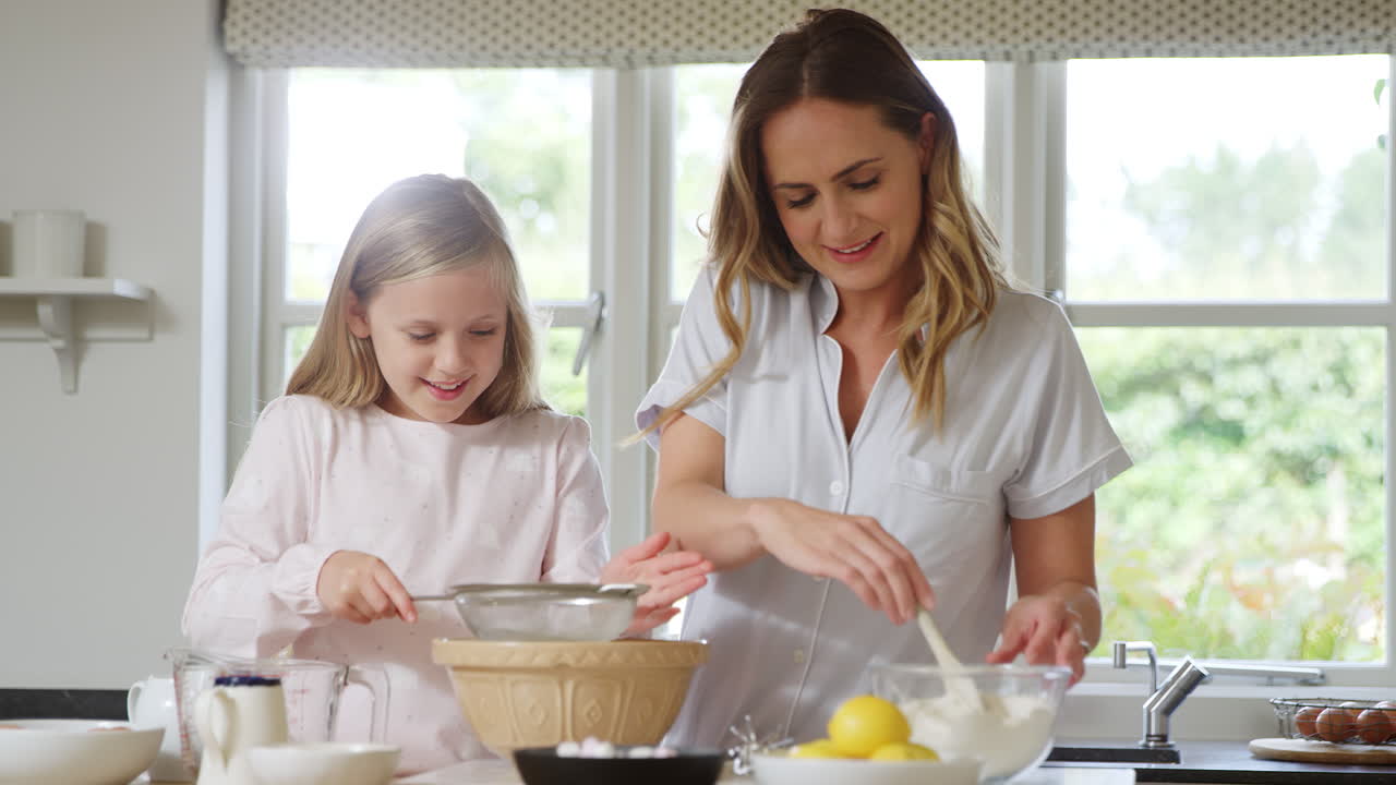 Mother And Daughter Wearing Pyjamas Baking And Making Pancakes In Kitchen At Home Together