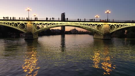 Low-fly-in-Guadalquivir-river,-passing-under-triana-bridge,-reveal-shot-of-pelli-sevilla-tower-during-sunset-dawn-in-Seville,-Spain