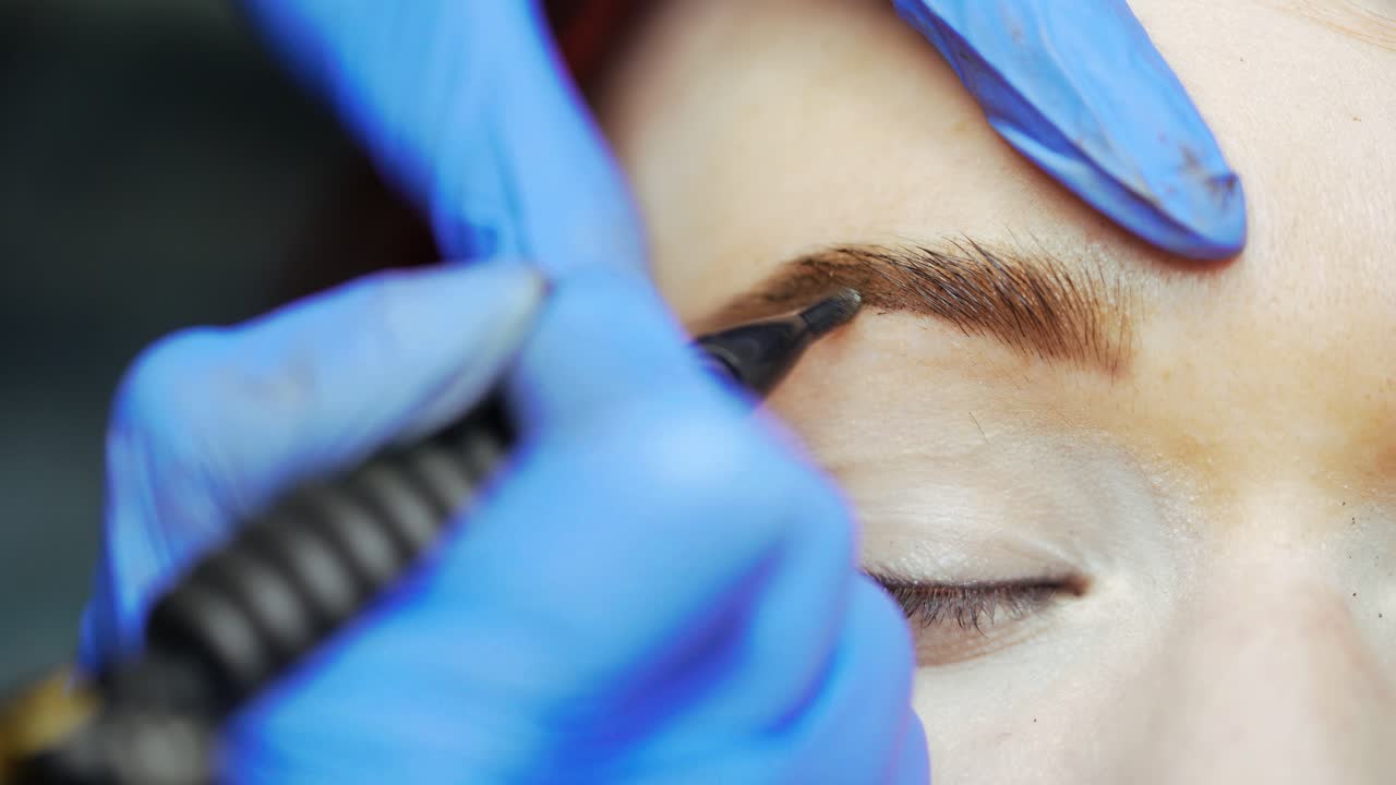 A beauty master is drawing the shape of the eyebrows using a thin needle for applying tattoo in the cosmetology. Close-up