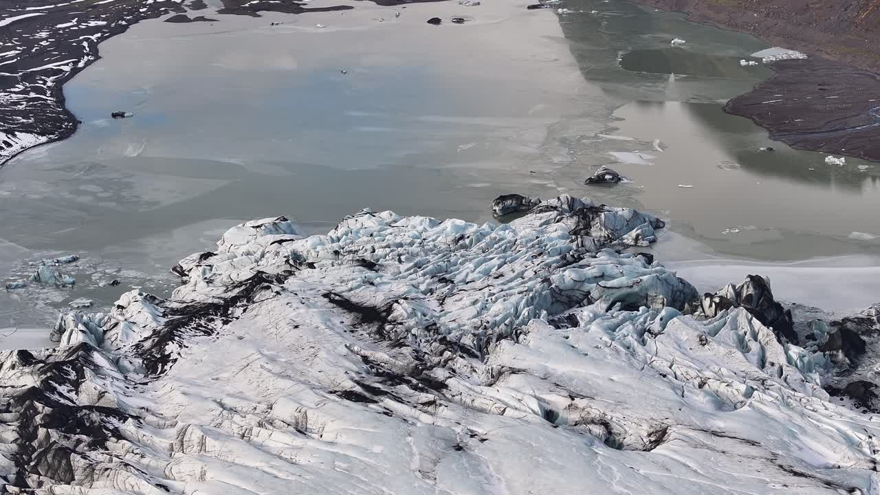 Aerial view of the edge of Sólheimajökull glacier in Iceland, showing dramatic ice formations merging into a frozen lagoon with floating icebergs.
