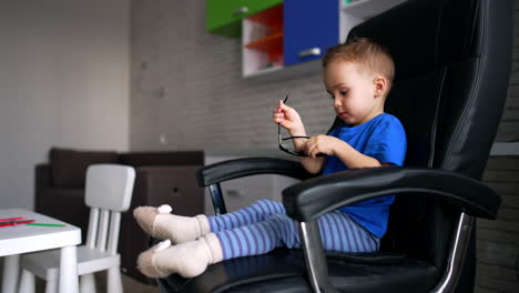 charming caucasian toddler sitting in comfortable chair indoors. baby boy putting on big sunglasses.