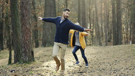 padre feliz caucásico y su pequeño hijo corriendo en el bosque con las manos abiertas imitando un avión volando