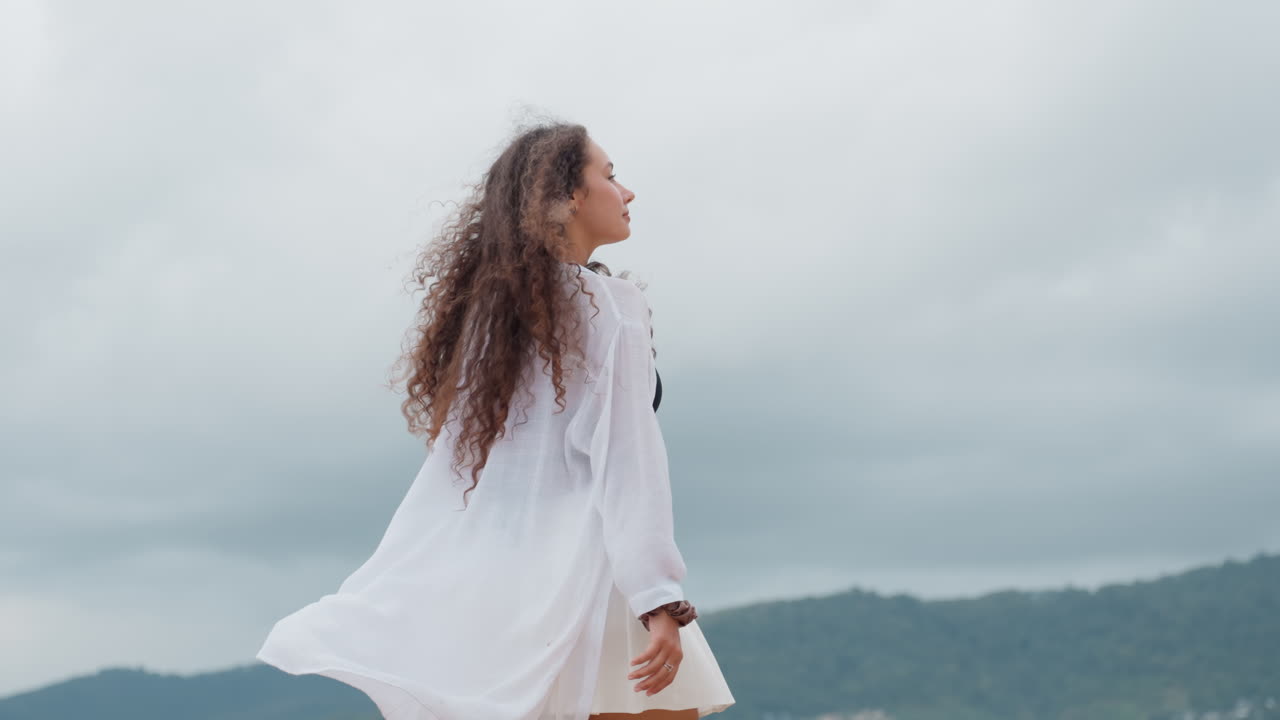 Donna di fronte all'orizzonte dell'isola sulla spiaggia, con la vestaglia che si gonfia al vento, silhouette di schiena contro il cielo nuvoloso, lento movimento di rotazione, postura contemplativa, montagne lontane, quiete meditativa, atmosfera costiera cinematografica.