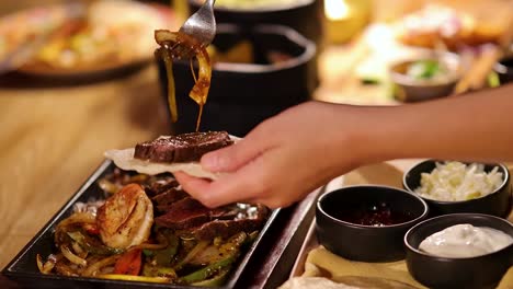 a person prepares a steak and shrimp fajita using a hot skillet, adding grilled vegetables and toppings under warm, ambient restaurant lighting