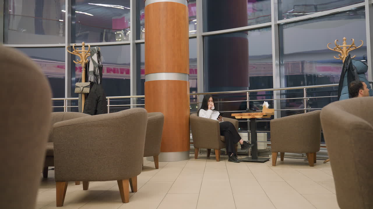 businesswoman sips drink then places it on table while browsing phone in stylish lounge with large windows, coat stands, soft chairs, and evening reflections creating warm modern indoor atmosphere