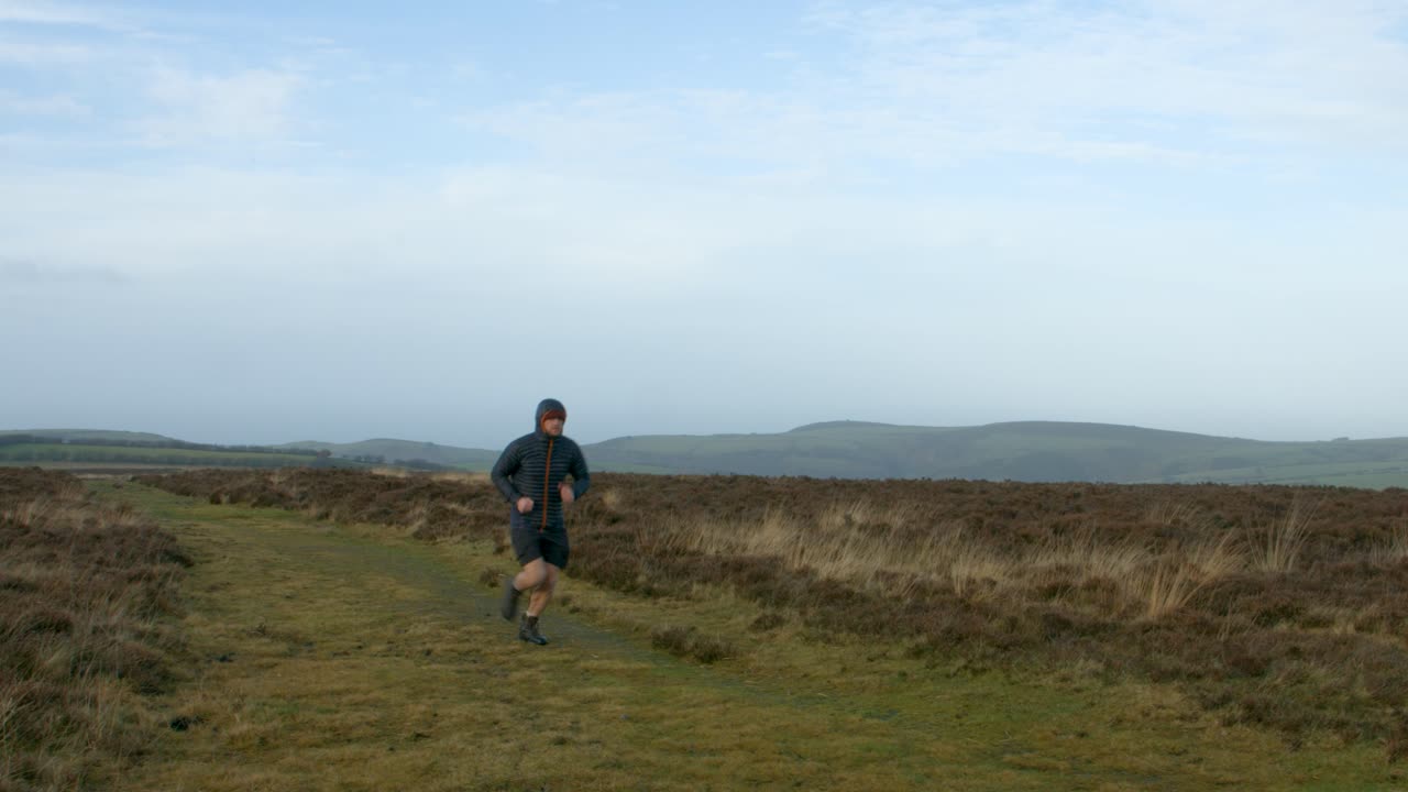 atleta de fitness masculino runner ejecutando entrenamiento en pantalones cortos y abrigo en condiciones de frío sobre páramos campiña exmoor uk 4k