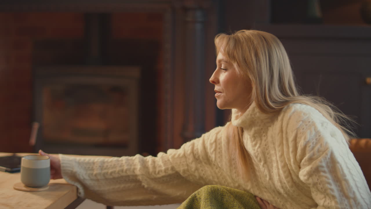 Woman At Home In Lounge With Cosy Fire With Hot Drink