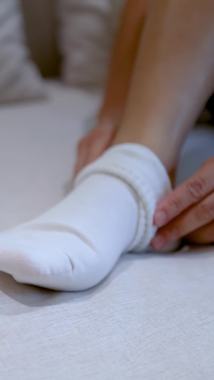 A woman sits on a bed and pulls on white cotton socks during morning routine.