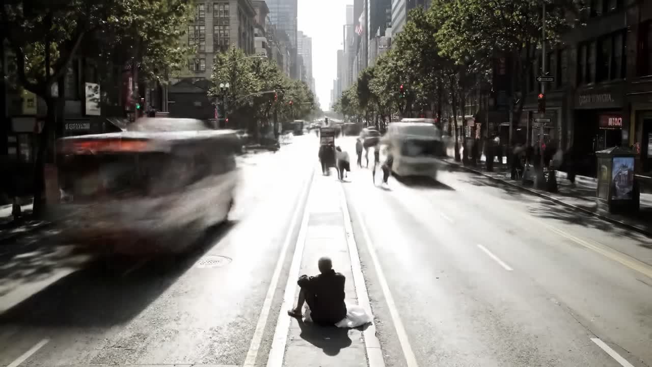 A bustling city street shows pedestrians moving along the sidewalk while a person sits on the dividing strip. Tall buildings reflect sunlight and create a vibrant urban atmosphere.