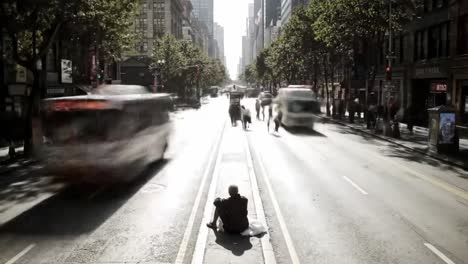 a bustling city street shows pedestrians moving along the sidewalk while a person sits on the dividing strip. tall buildings reflect sunlight and create a vibrant urban atmosphere.