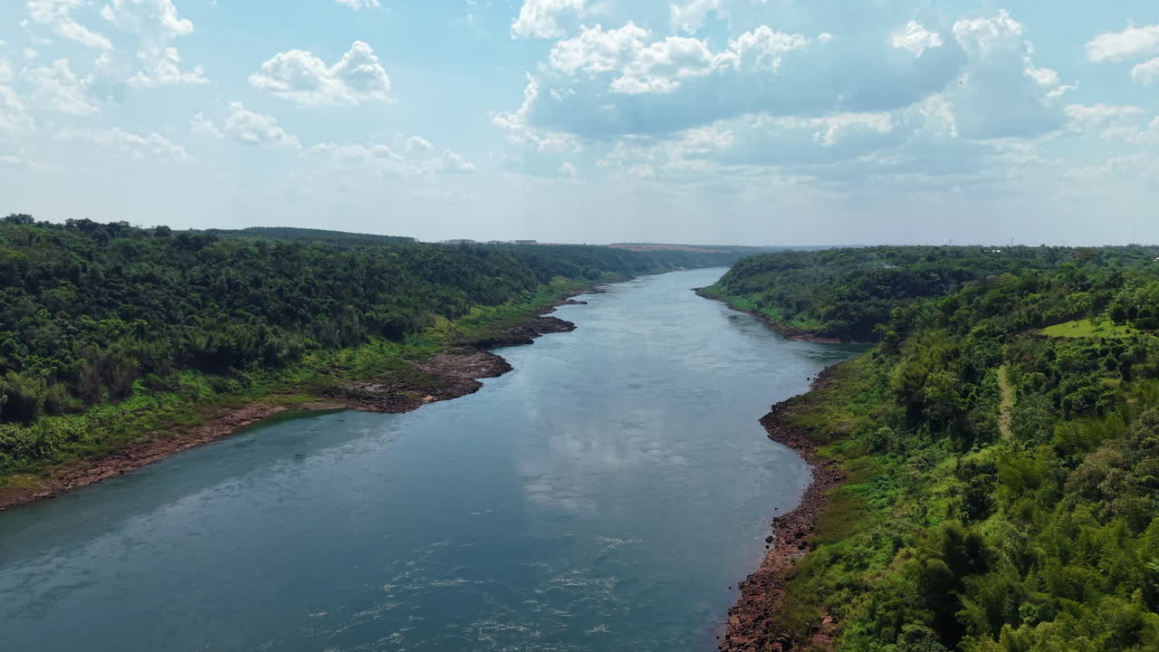 Smooth aerial glide above the Paraná River, showcasing vast tropical vegetation under sunny sky with cumulus clouds