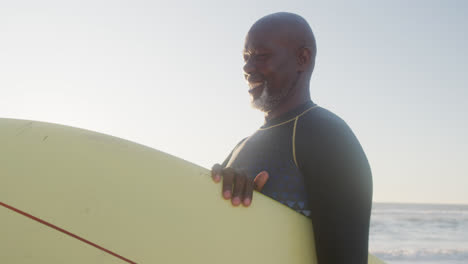 Happy-senior-african-american-man-walking-and-holding-surfboard-at-beach,-in-slow-motion