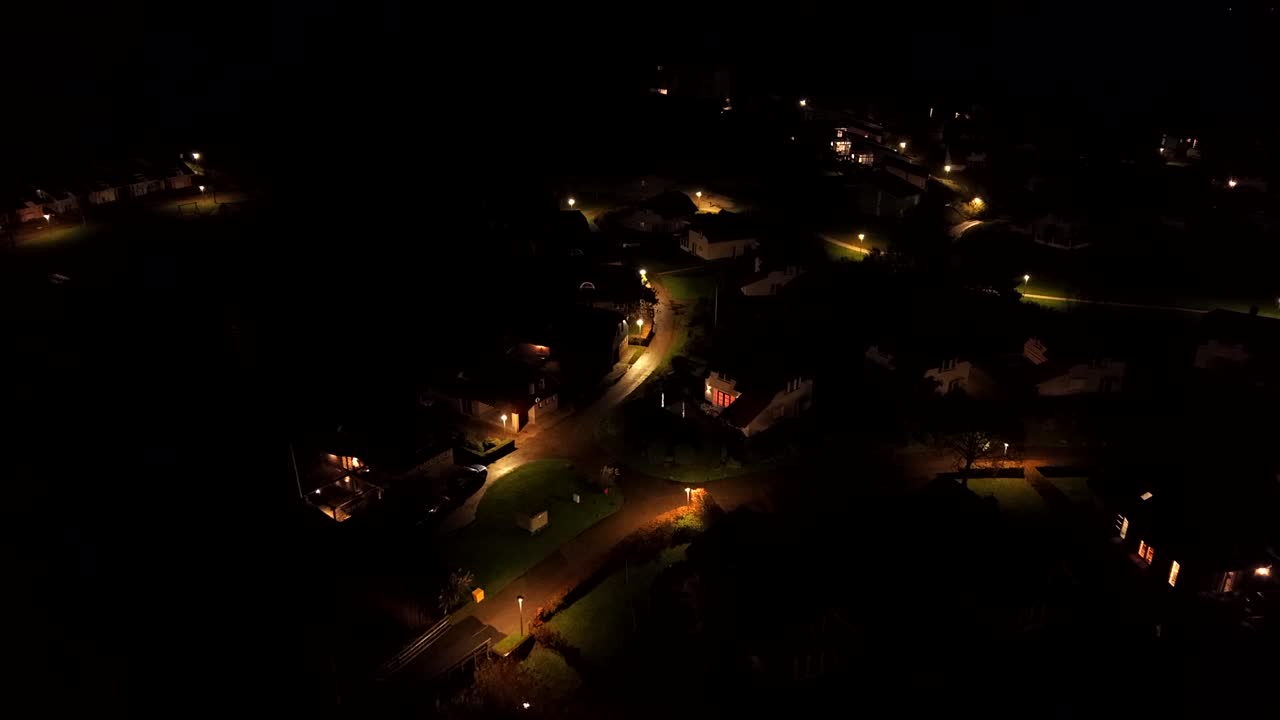 Aerial night view of an American suburban neighborhood illuminated by warm streetlights, showing quiet streets, houses and a peaceful residential atmosphere. Birds Eye flight
