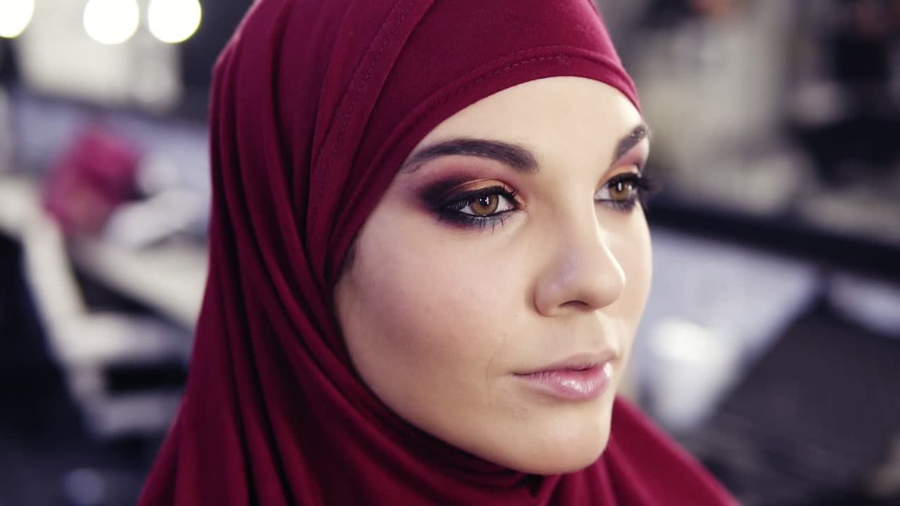 imágenes en cámara lenta de una hermosa chica caucásica con un hijab de chiffon púrpura en la cabeza recibiendo los toques finales de su maquillaje para los ojos.