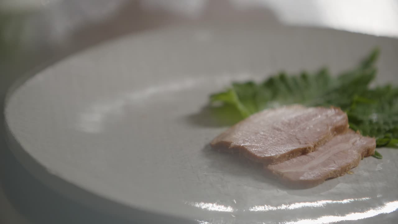 A close-up view of a professional chef carefully plating a gourmet dish using tongs. The setting is a modern kitchen, and the chef's attention to detail highlights high-end culinary presentation.
