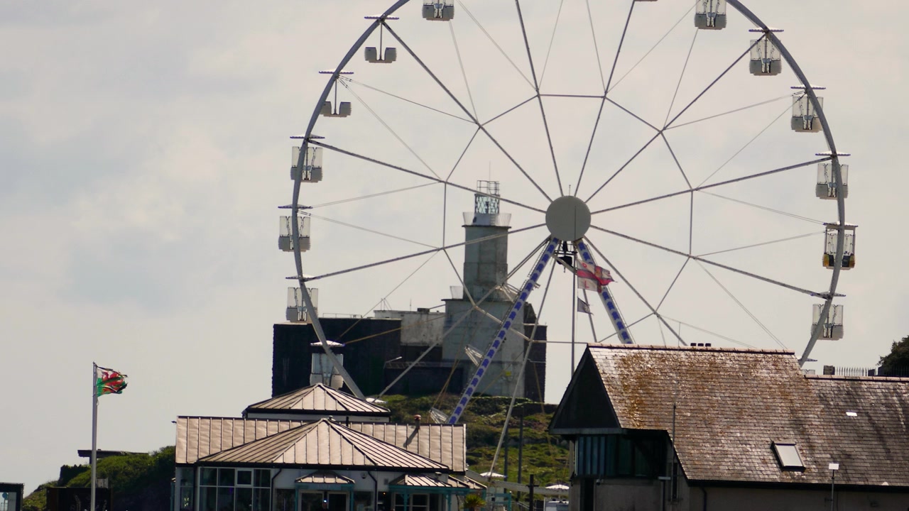 Ett stort pariserhjul mot en molnig himmel, inramat av hustak och en walesisk flagga som vajar i vinden i Mumbles, Wales