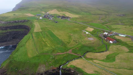 Toma-Aérea-De-Una-Gaviota-Volando-Sobre-El-Pueblo-De-Gasadalur-Y-Sus-Tierras-De-Cultivo-Cerca-De-La-Cascada-De-Mulafossur