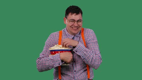 un hombre ve una película divertida, ríe y come palomitas de maíz de un cubo grande. retrato de un hombre con gafas, en una camisa con tirantes naranjas en el estudio en una pantalla verde. cámara lenta.
