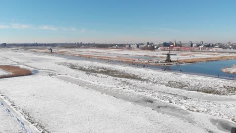 Dutch-locals-ice-skating-on-frozen-canal-near-old-windmill,-winter-aerial-view