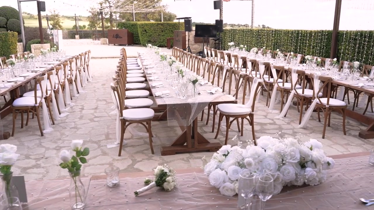 File di tavoli sono apparecchiati per un grande evento all'aperto. Ogni tavolo ha piatti, bicchieri e fiori posizionati su di esso. L'ambiente è tranquillo con uno sfondo verde