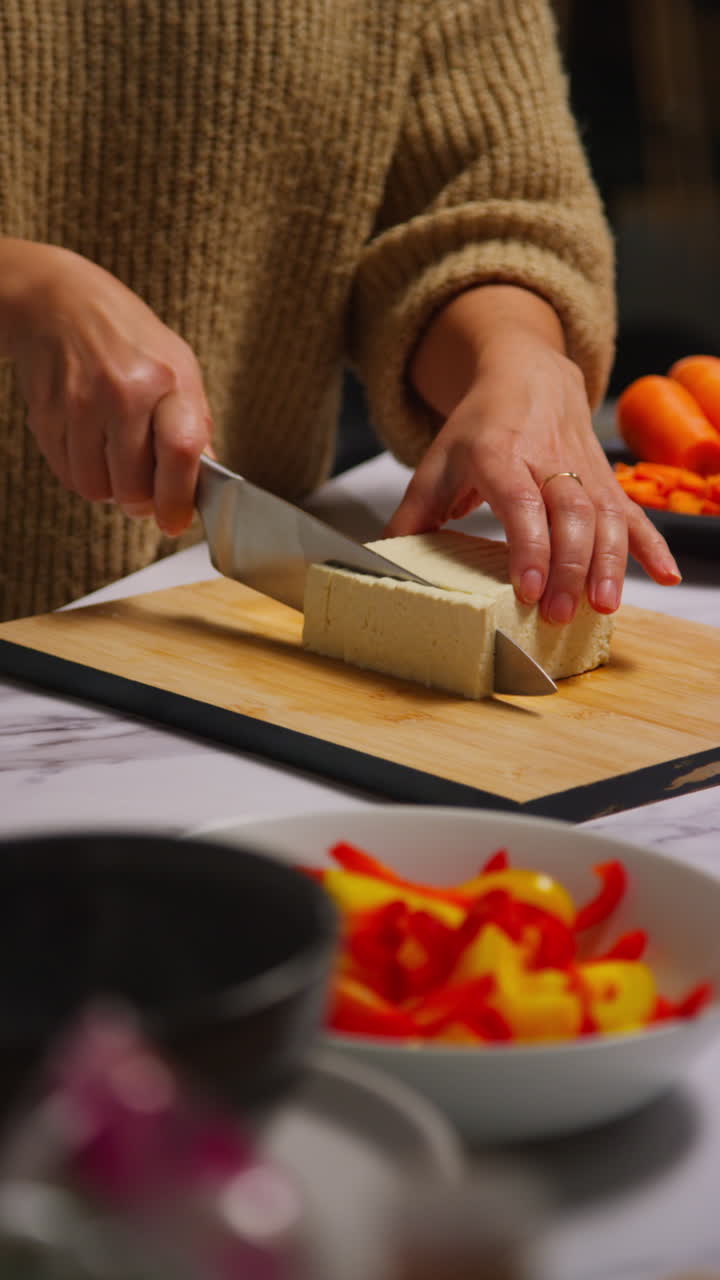 Primo piano verticale di una donna a casa in cucina che prepara un pasto vegetariano o vegano sano, mentre taglia il tofu su una tavola con un coltello 1