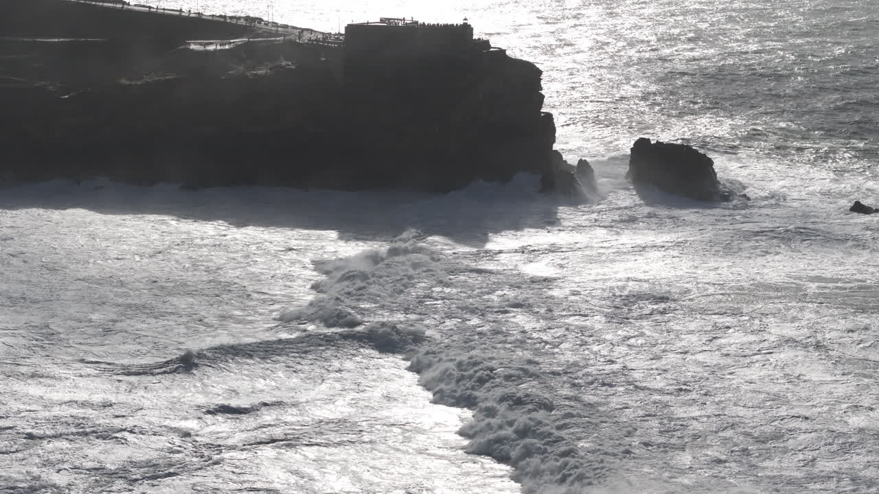 Aerial drone wide and high helicopter style establishing shot of Nazaré, Portugal, Europe. Farol da Nazaré lighthouse and lines of big waves rolling into shore