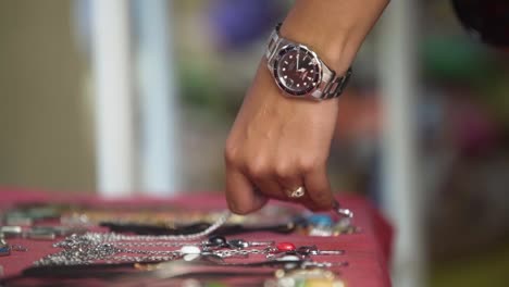 Primer-Plano-De-La-Mano-Femenina-Eligiendo-Joyas-Del-Mercado-De-Pulgas-De-La-Calle-Mano-Con-Reloj-Y-Anillo-De-Plata,-Algunas-Pulseras-En-La-Mesa