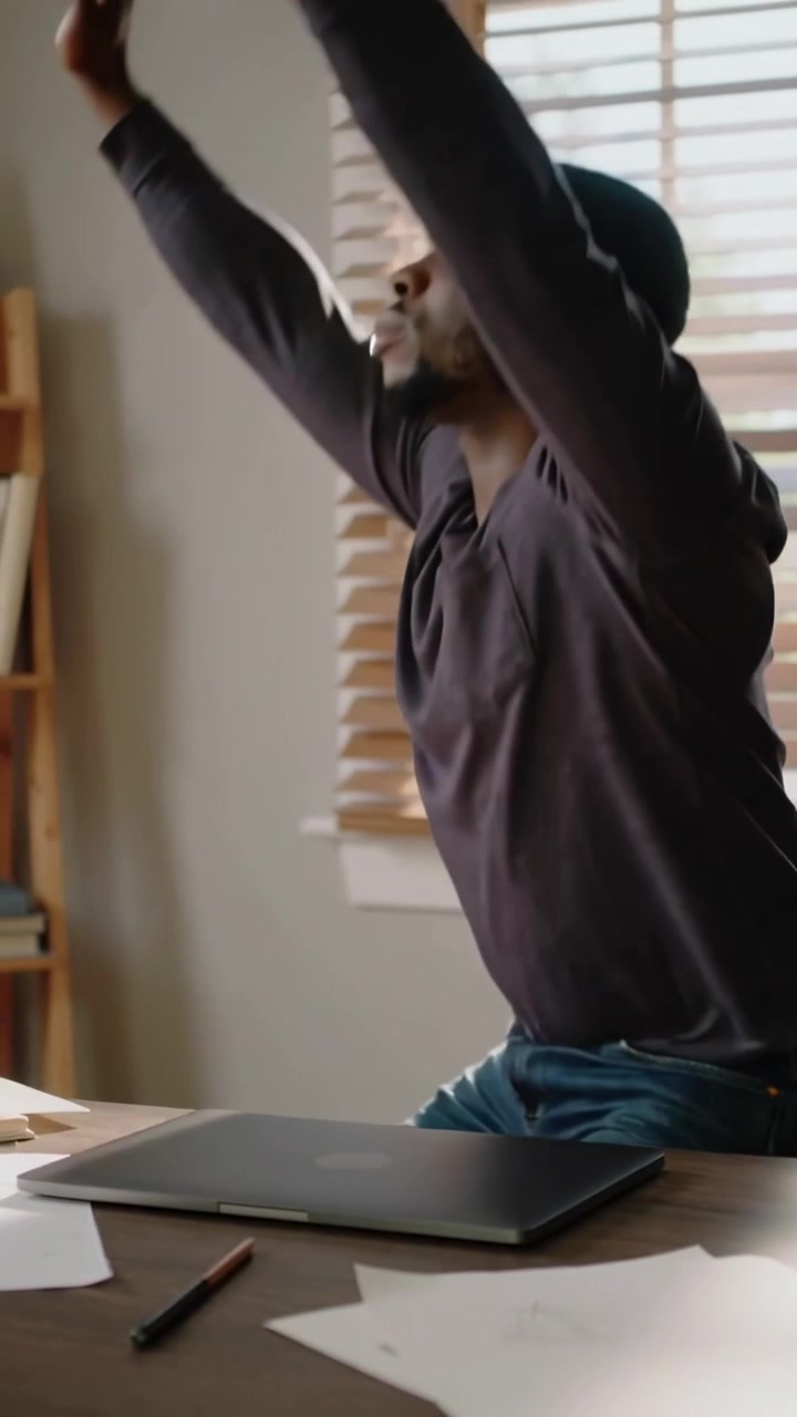 Video vertical: Hombre de camisa oscura haciendo una pausa estirándose en el escritorio de casa para un descanso con laptop, espacio para texto