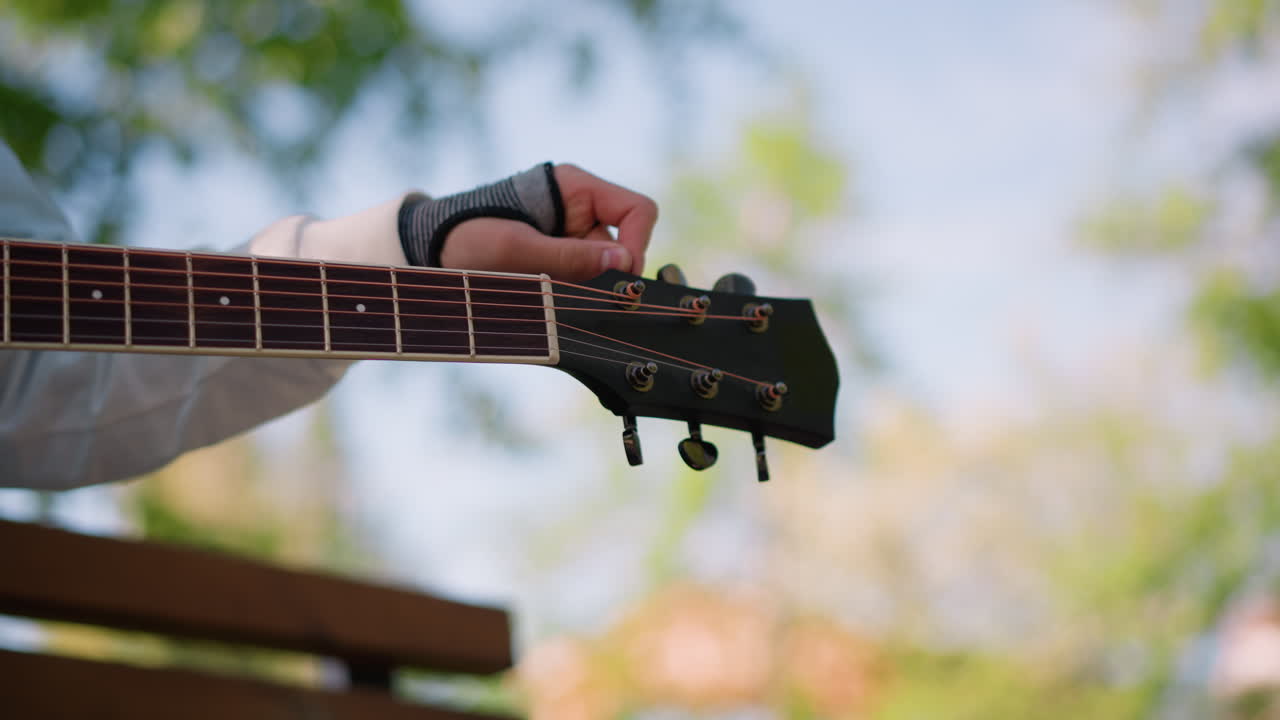 Escena relajada al aire libre con guitarra y toldo verde. Una mano ajusta la guitarra en el exterior bajo una cubierta verde frondosa. Ambiente informal al aire libre donde alguien está afinando y tocando la guitarra acústica.