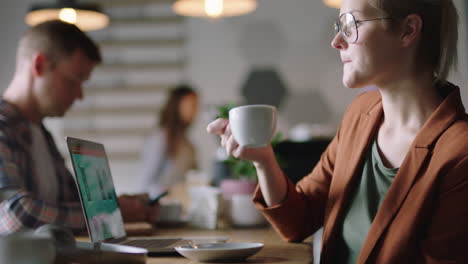 young caucasian business woman engineer using laptop computer in cafe developing mobile software drinking coffee reading online document wearing glasses enjoying relaxed restaurant