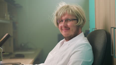 vista en primer plano de un técnico con una peluca rubia y gafas, sonriendo a la cámara mientras está sentado en un escritorio de laboratorio con un microscopio