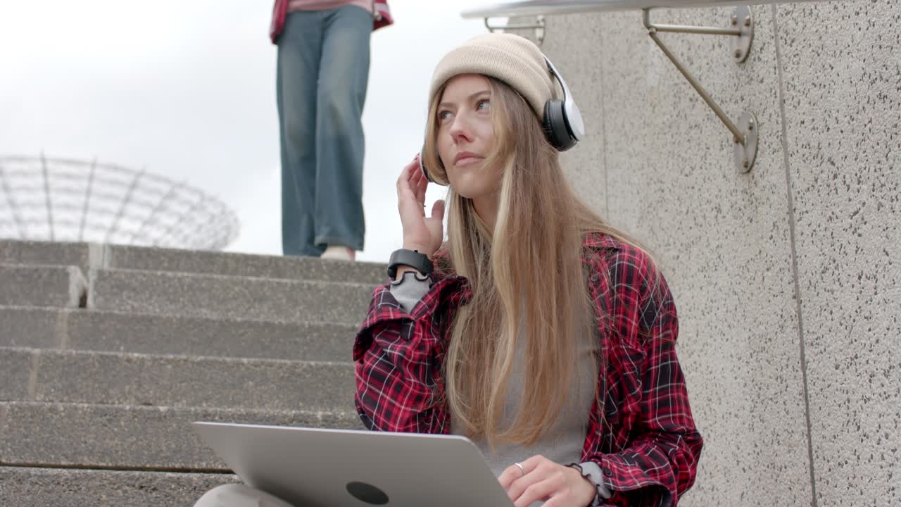 Studente che digita al computer portatile sui gradini, si ferma, prende le cuffie e ascolta la musica